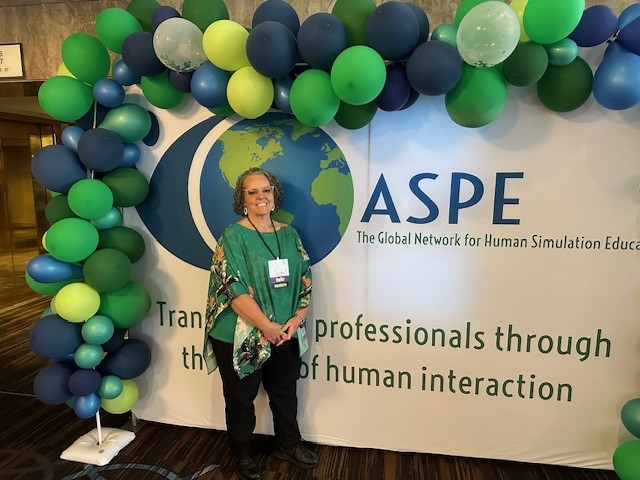 One sentence alt text for this image: A smiling woman stands in front of a colorful balloon arch and ASPE conference banner that reads “Transforming professionals through the power of human interaction.”