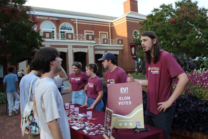 Students in maroon “Elon Votes!” shirts engage passersby at an outdoor voter registration table in front of the Moseley Center.