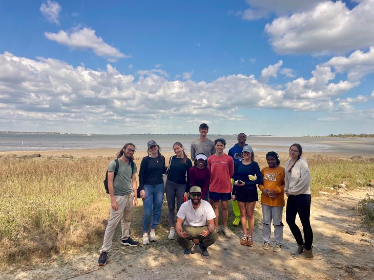 A group of students poses together on a sunny shoreline with marsh grass and open water in the background.