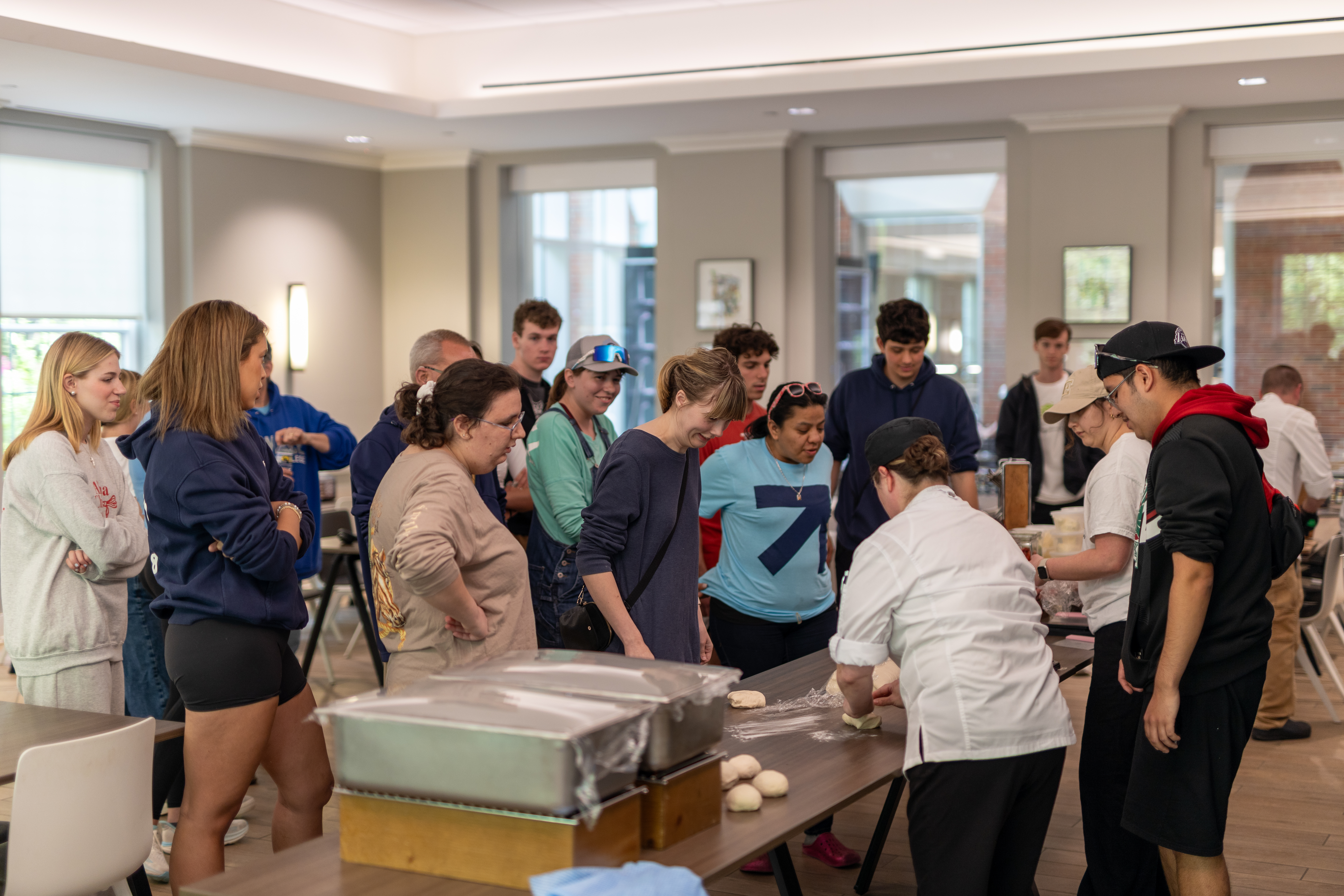  A chef demonstrates how to shape dough as a group of students and adults attentively watches during an indoor baking workshop.