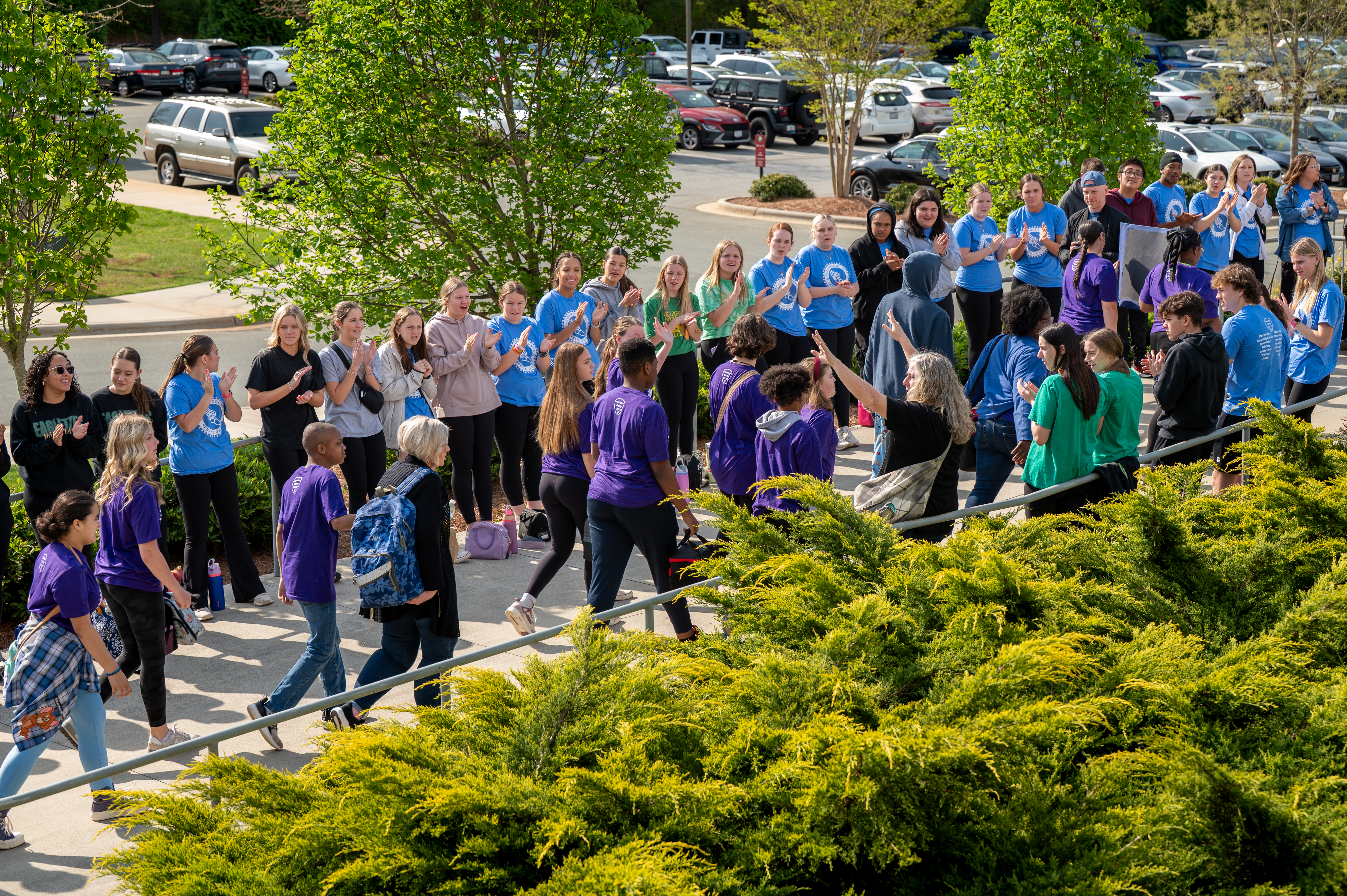 Students wearing colorful shirts cheer and applaud others walking between them in a festive outdoor gathering on a sunny day.