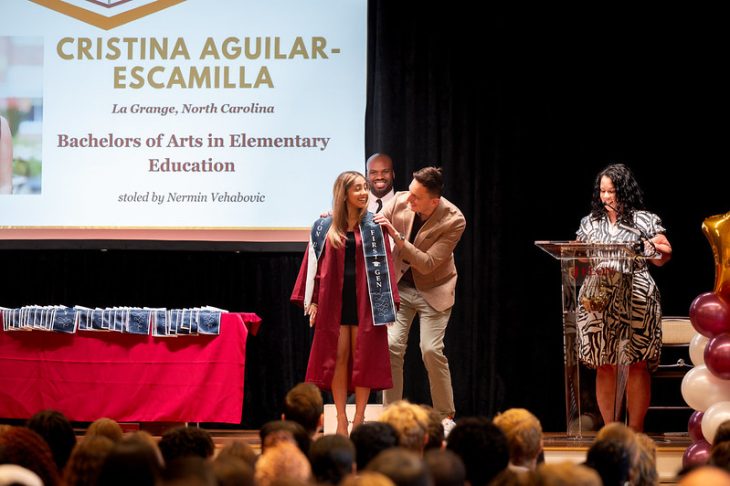 A smiling graduate in a maroon cap and gown receives a First-Gen stole on stage during a ceremony, with a projected slide displaying her name and degree in the background.