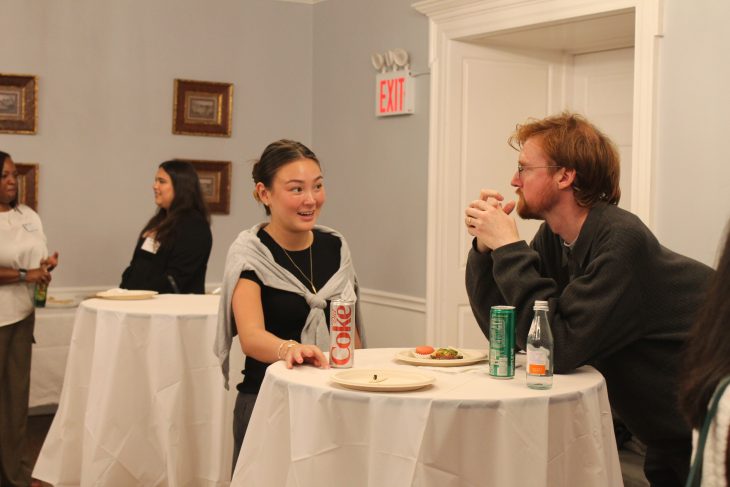 Two people smile and talk at a standing table during a casual social event, with drinks and plates of food in front of them.