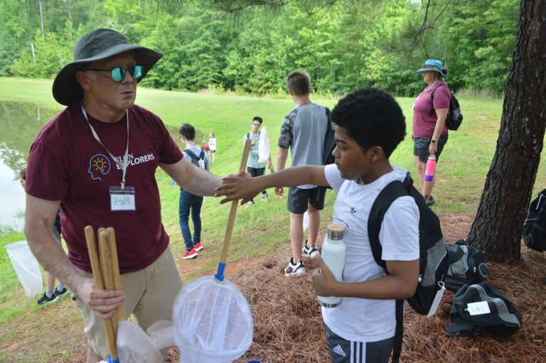 A boy reaches out to take a net from an adult leader wearing an “Elon Explorers” shirt, while other participants explore near a pond in the background.