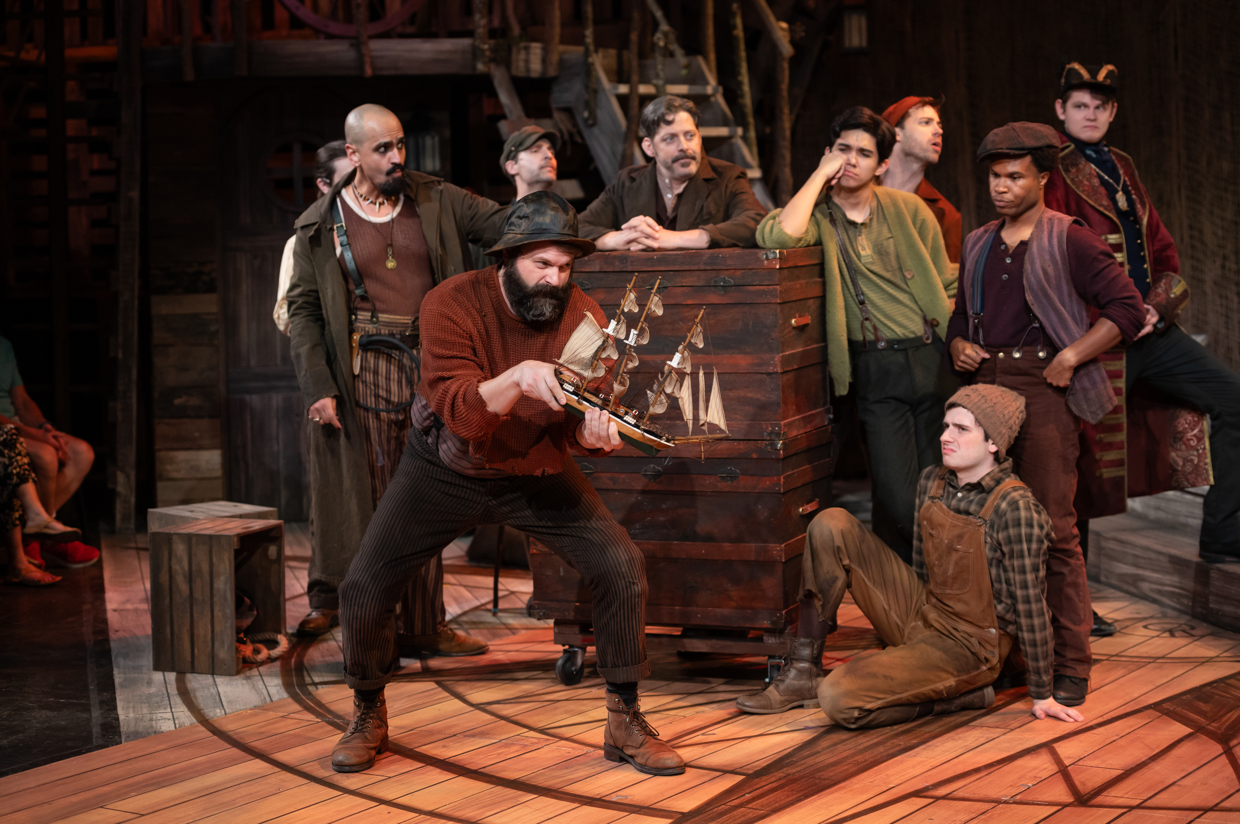 A group of actors in period costumes perform a dramatic scene on stage, with one actor holding a model ship while others watch intently.