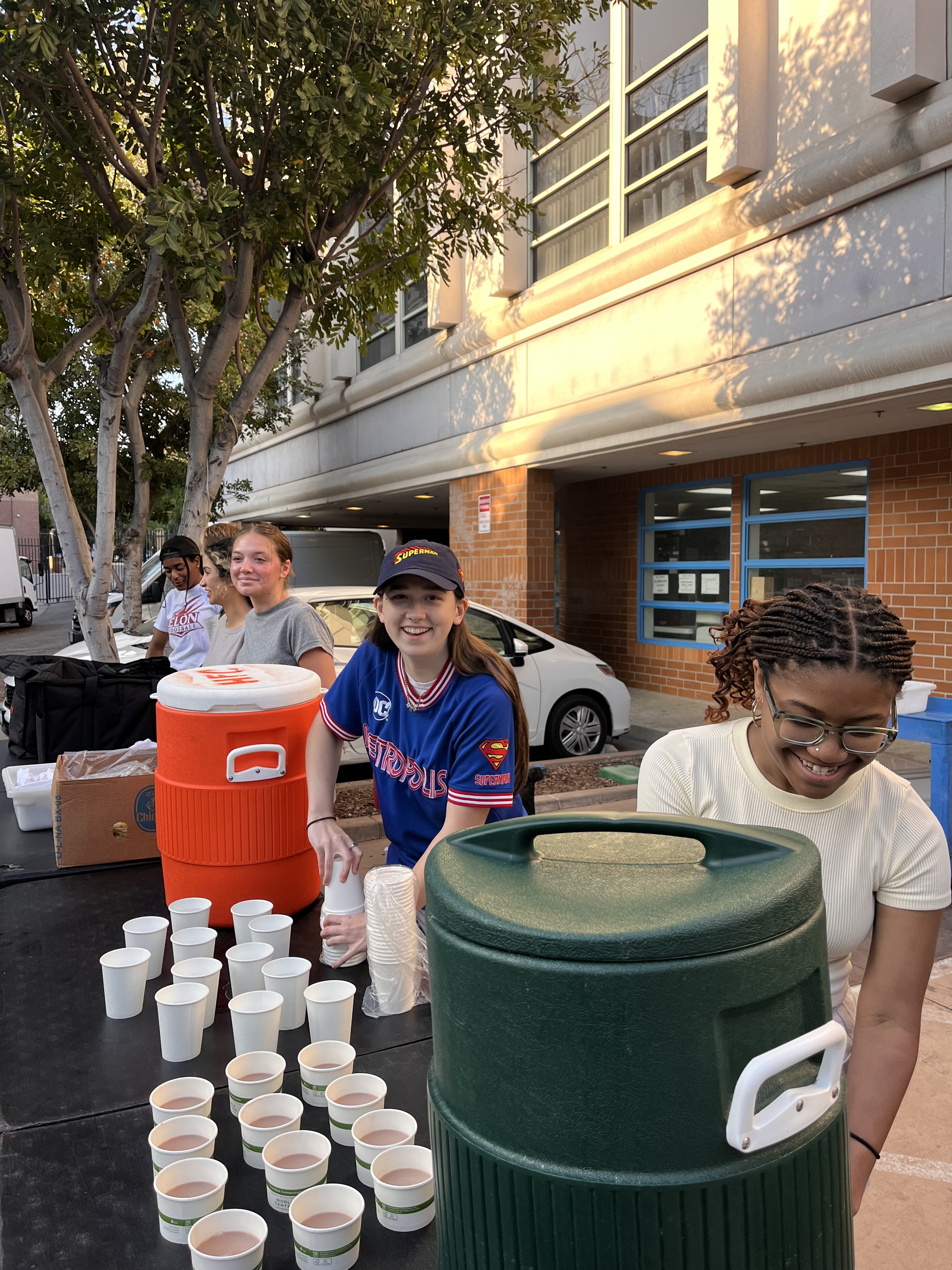 Three volunteers happily serve drinks from large coolers outdoors, with cups of juice lined up on the table in front of them.