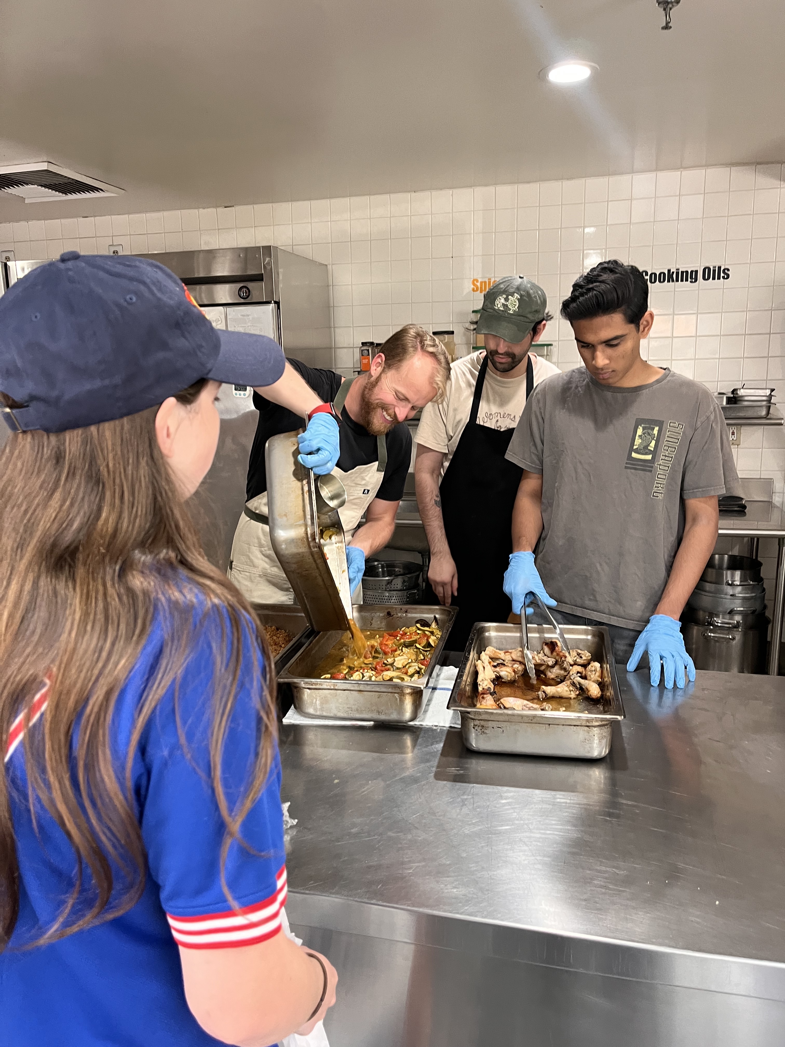 Volunteers in a kitchen prepare and serve trays of food, as one person pours sauce over cooked vegetables.