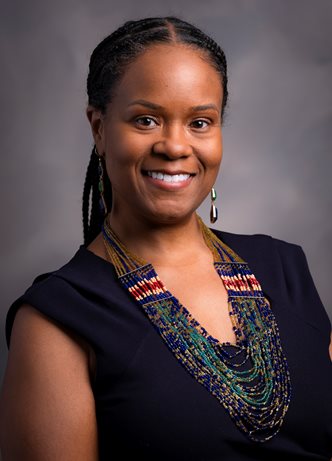 The photo shows a woman posing confidently in front of a neutral gray background. She has a warm, friendly smile and is wearing a dark, sleeveless dress. Her hair is styled in neat braids pulled back. She's adorned with a vibrant, multi-strand beaded necklace that features a mix of blue, green, red, and gold tones, and she's also wearing dangling earrings. The lighting and background suggest this is a professional portrait, possibly for business or academic use.
