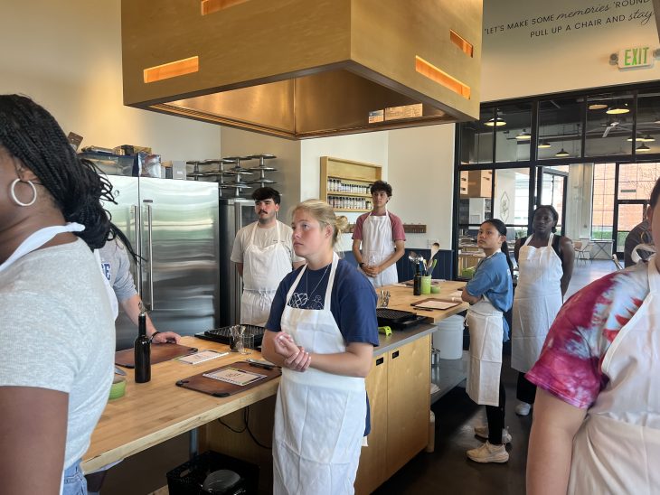 A group of students wearing aprons listens attentively during a cooking class in a modern kitchen setting.