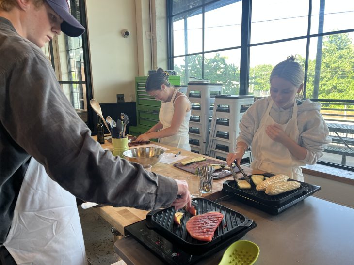 Three students in aprons prepare food at a kitchen counter, grilling vegetables and slicing ingredients.
