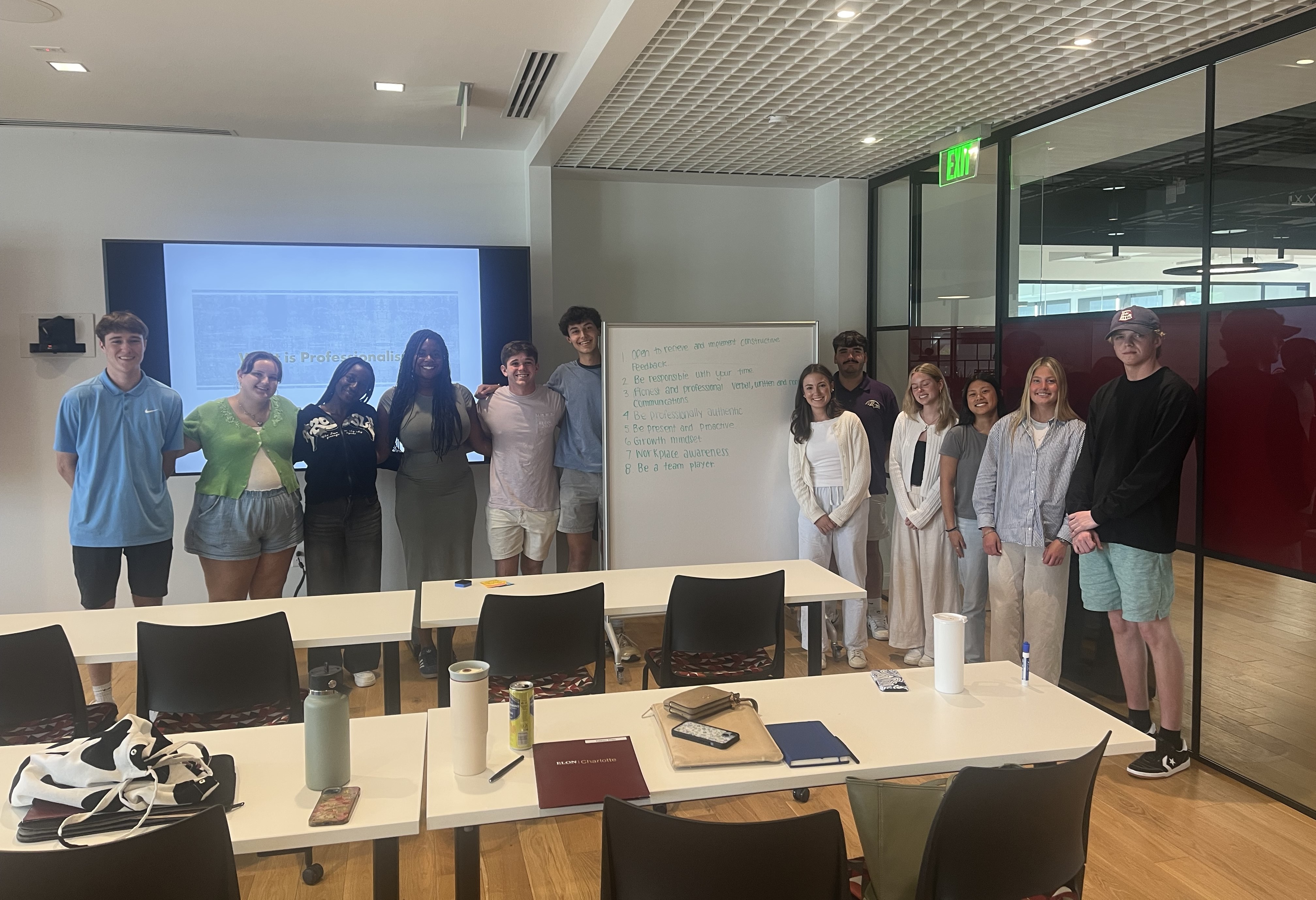 A group of students and instructors pose in front of a whiteboard and screen inside a classroom, smiling after a presentation.