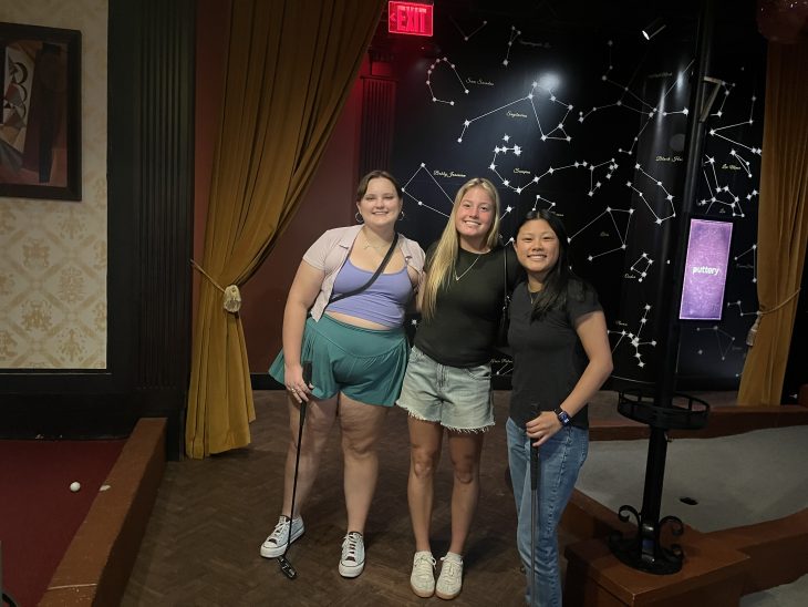 Three students with mini-golf clubs pose together in front of a constellation-themed wall at an indoor entertainment venue.