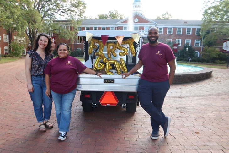 Three people smile while standing behind a decorated cart with gold balloons spelling “FIRST GEN” on Elon University’s campus near a fountain.