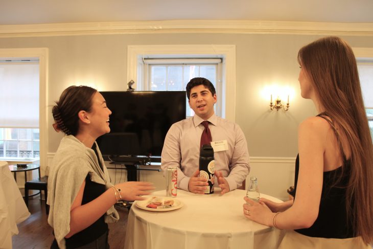 Two women and one man talk and laugh around a cocktail table with drinks and snacks during a casual networking event.