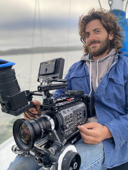 John Giacobbe holds a camera while sitting on a sailboat.