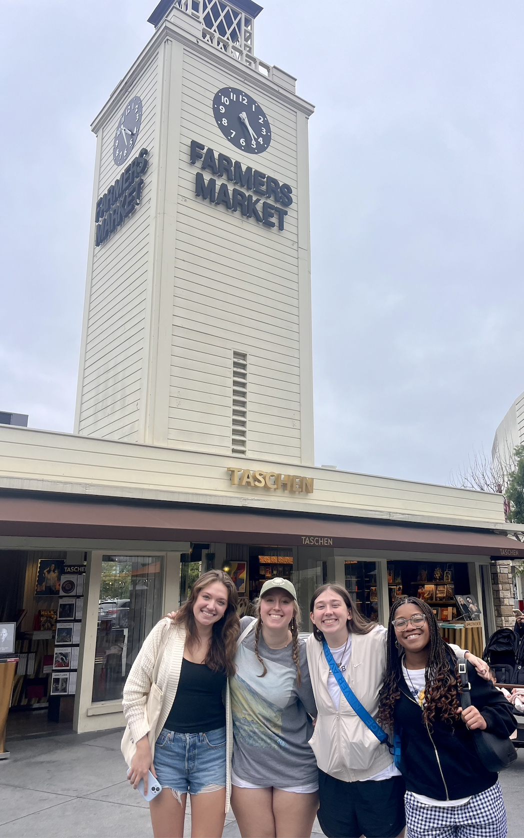 Four smiling friends pose arm-in-arm in front of the iconic Farmers Market clock tower in Los Angeles.