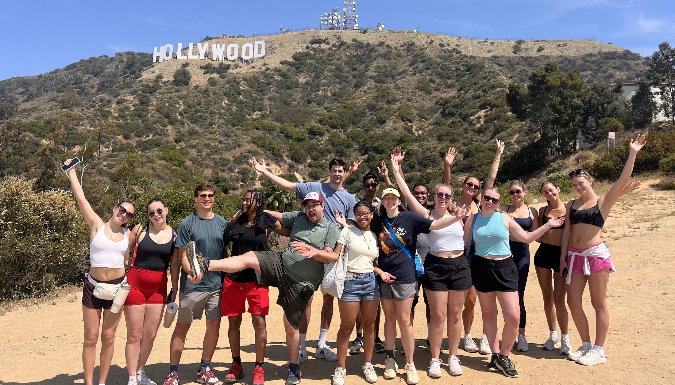 A group of smiling young adults poses with arms raised in front of the iconic Hollywood sign on a sunny hiking trail.