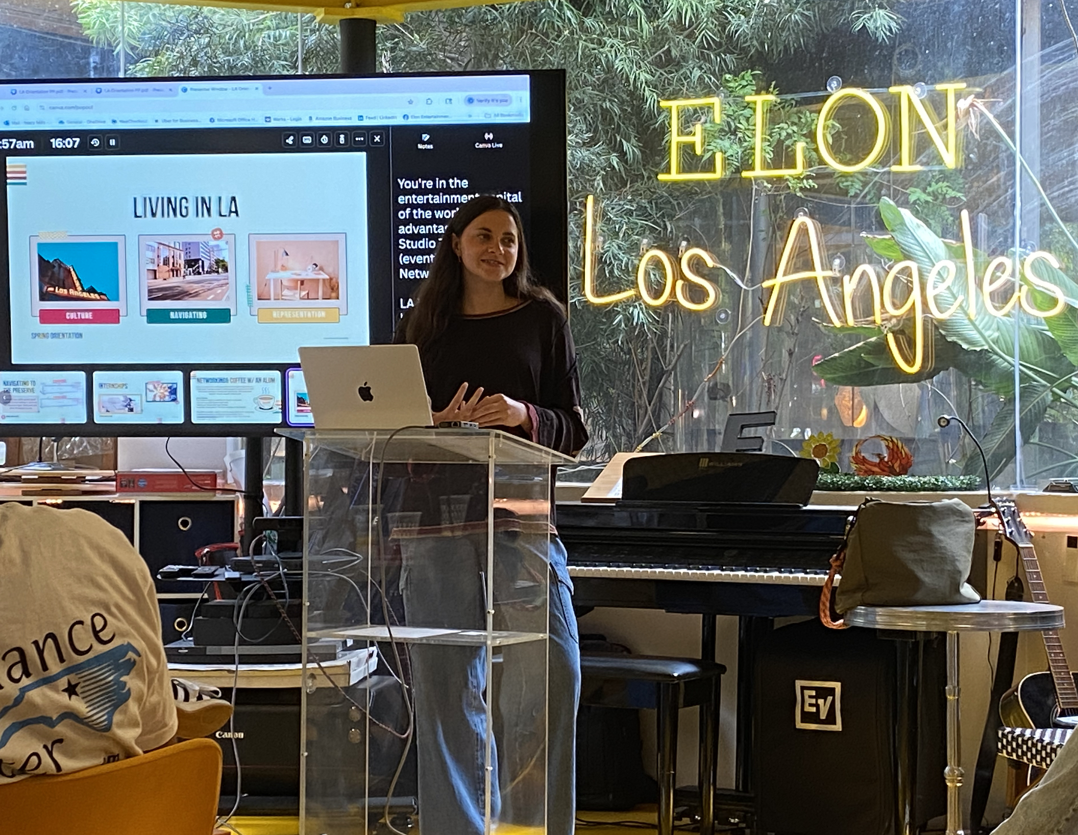 A woman stands at a clear podium giving a presentation about living in Los Angeles, with a neon “Elon Los Angeles” sign glowing behind her.