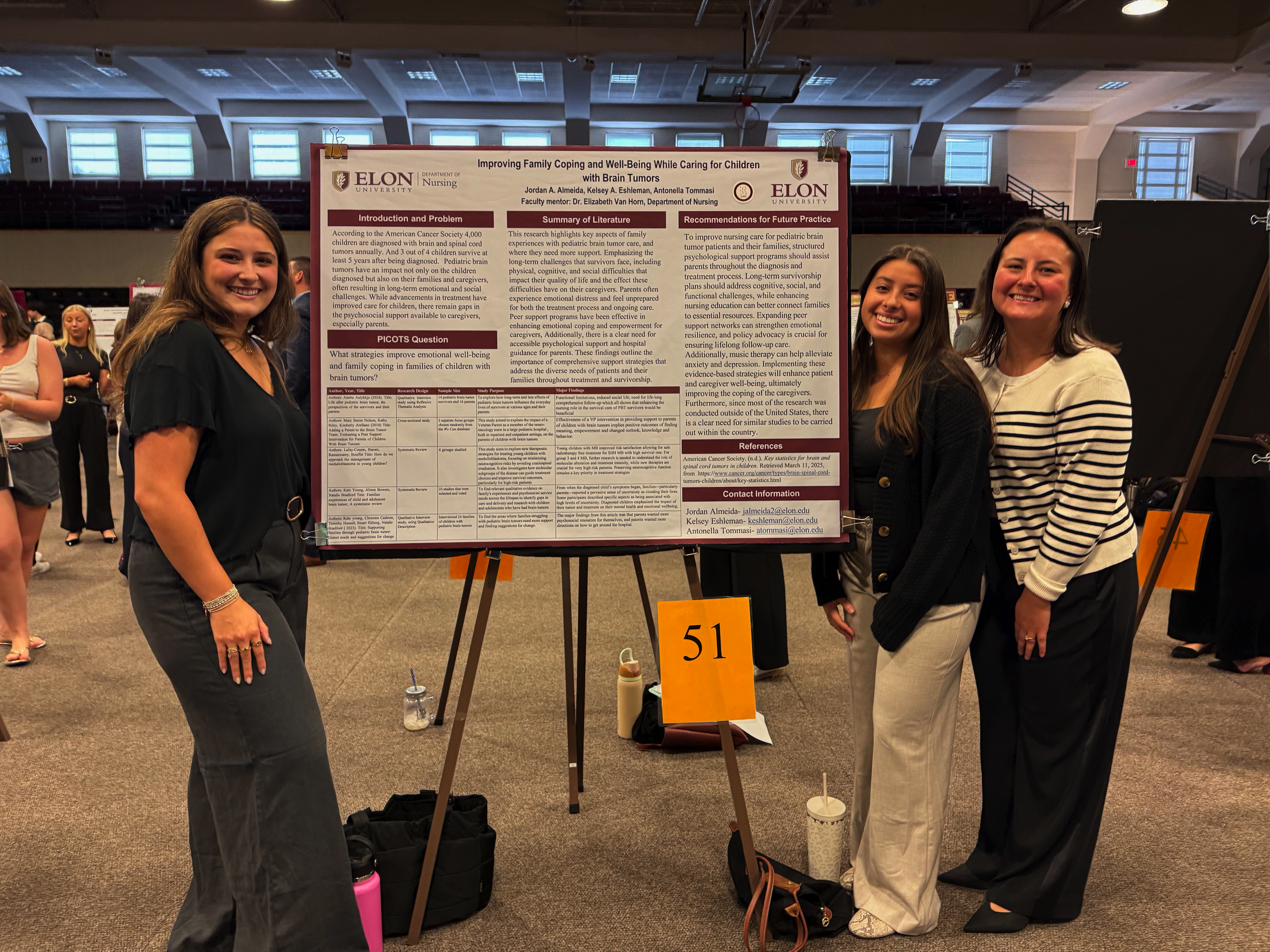 Three women smile and stand beside a large academic research poster on improving family coping for children with brain tumors at a campus presentation event.