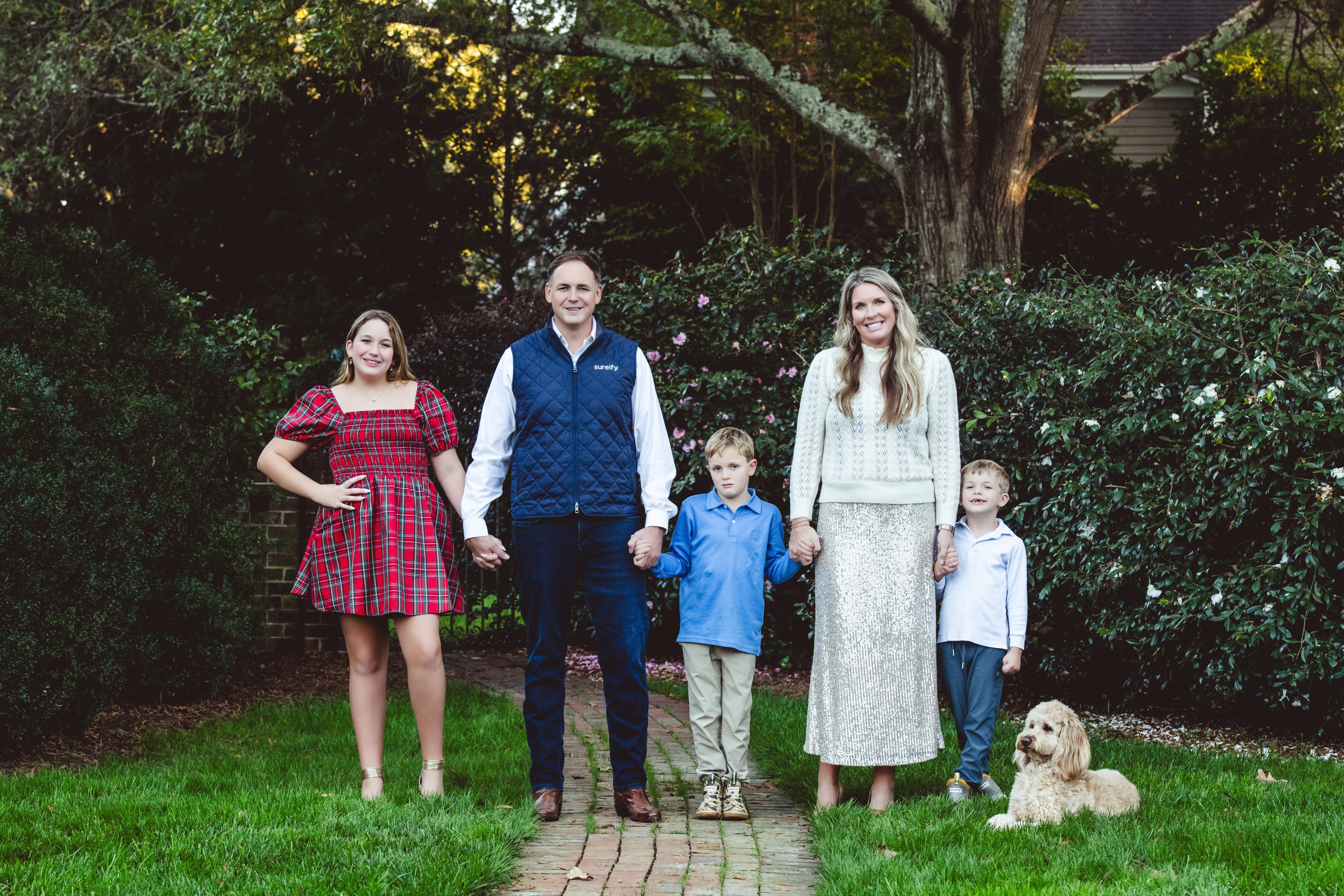 A family of five and their dog pose for a portrait outdoors on a brick path, surrounded by greenery and trees.