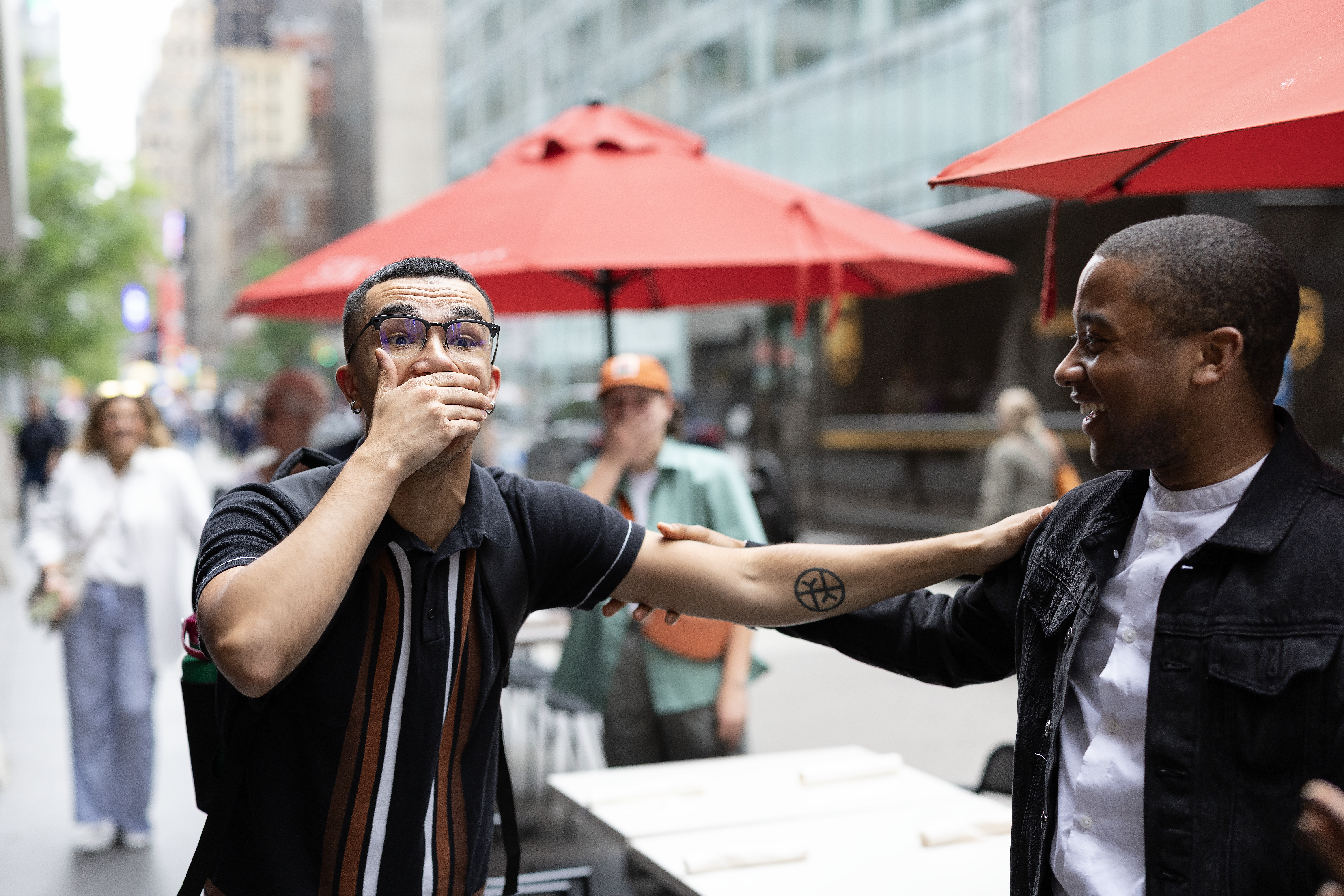 A surprised young man covers his mouth in shock as another man smiles and places a hand on his shoulder on a city sidewalk.