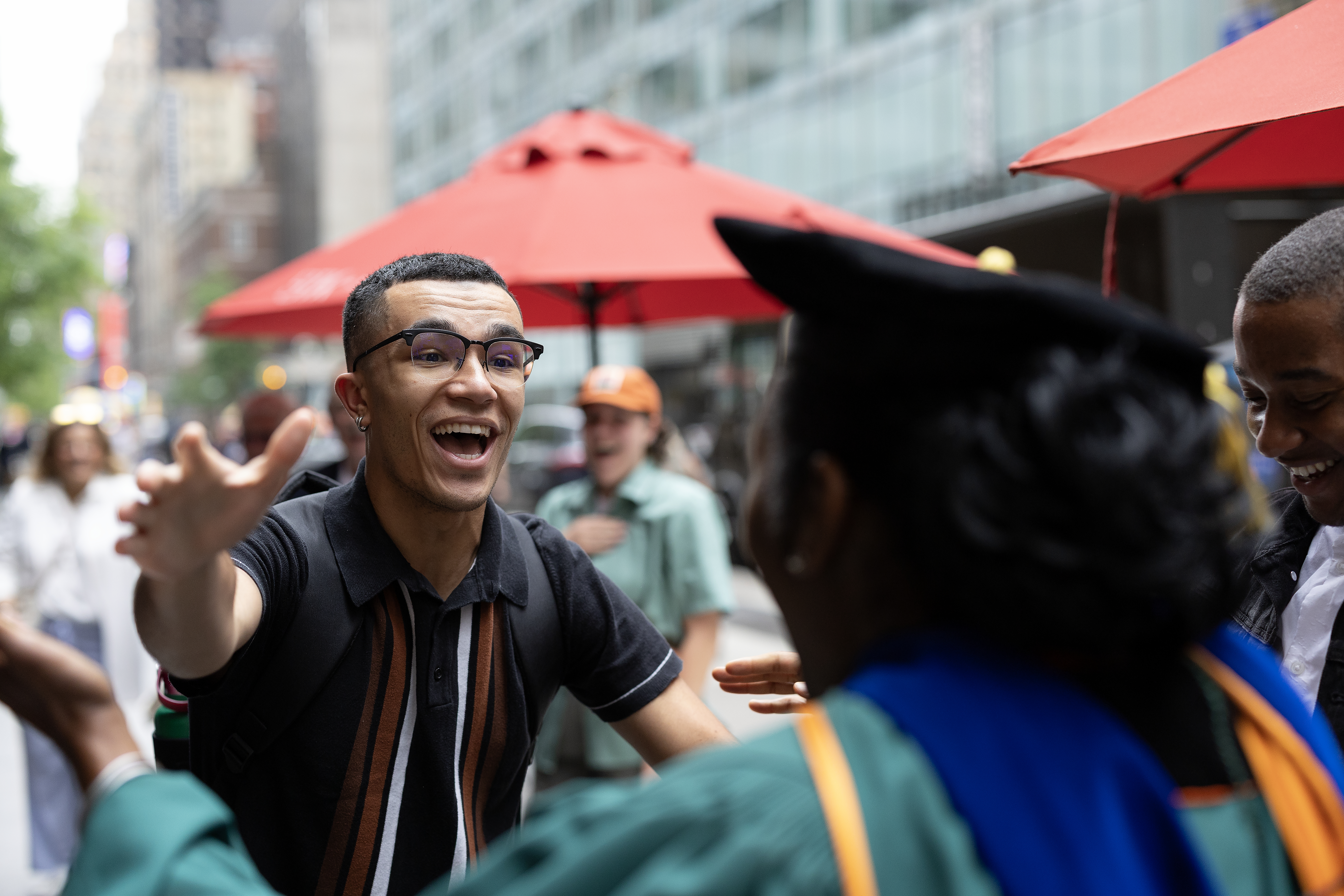 A joyful young man with outstretched arms greets someone in academic regalia on a city street lined with red umbrellas, surrounded by smiling onlookers.