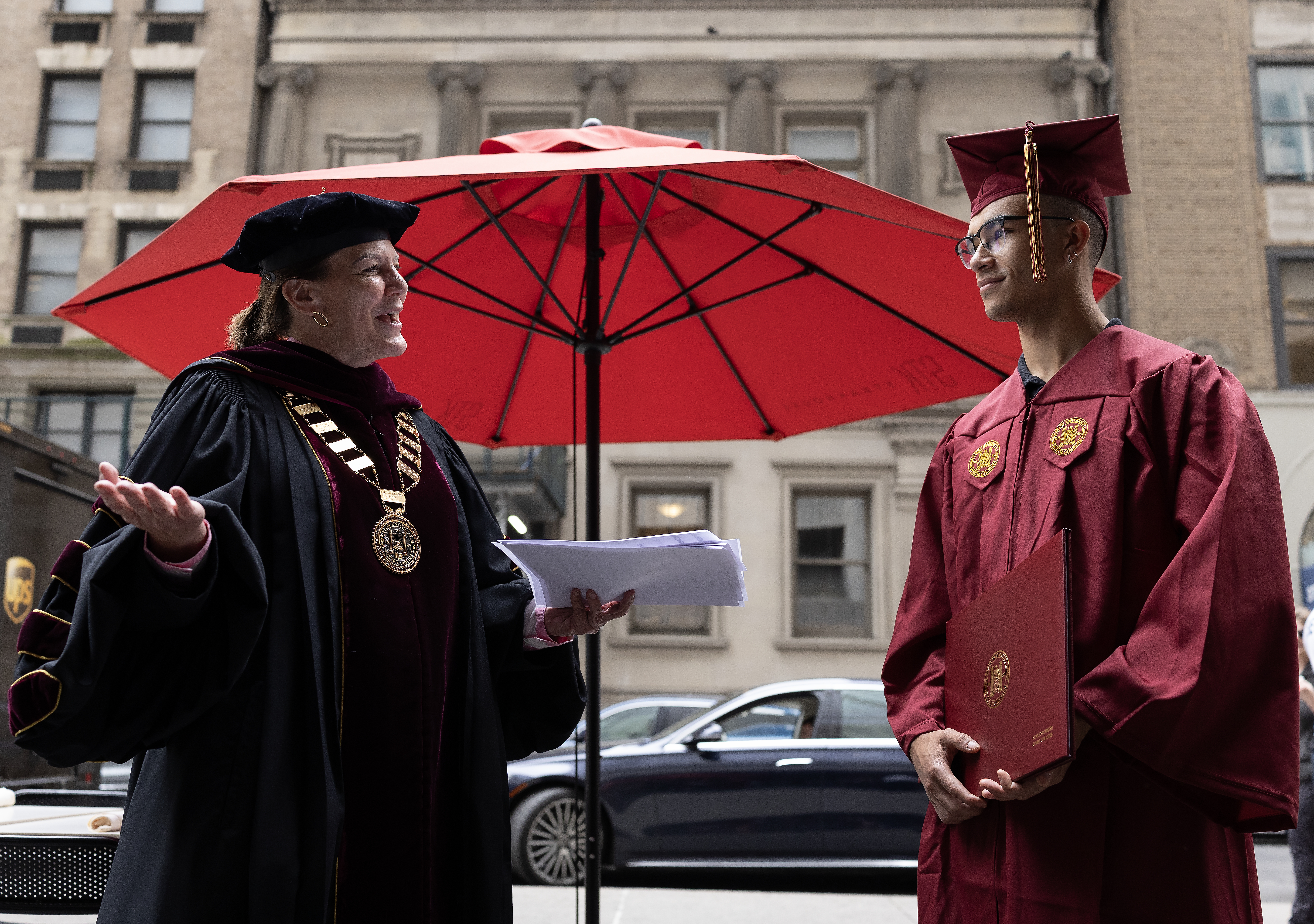 A university president in formal academic regalia smiles and hands a document to a graduate in a maroon cap and gown.
