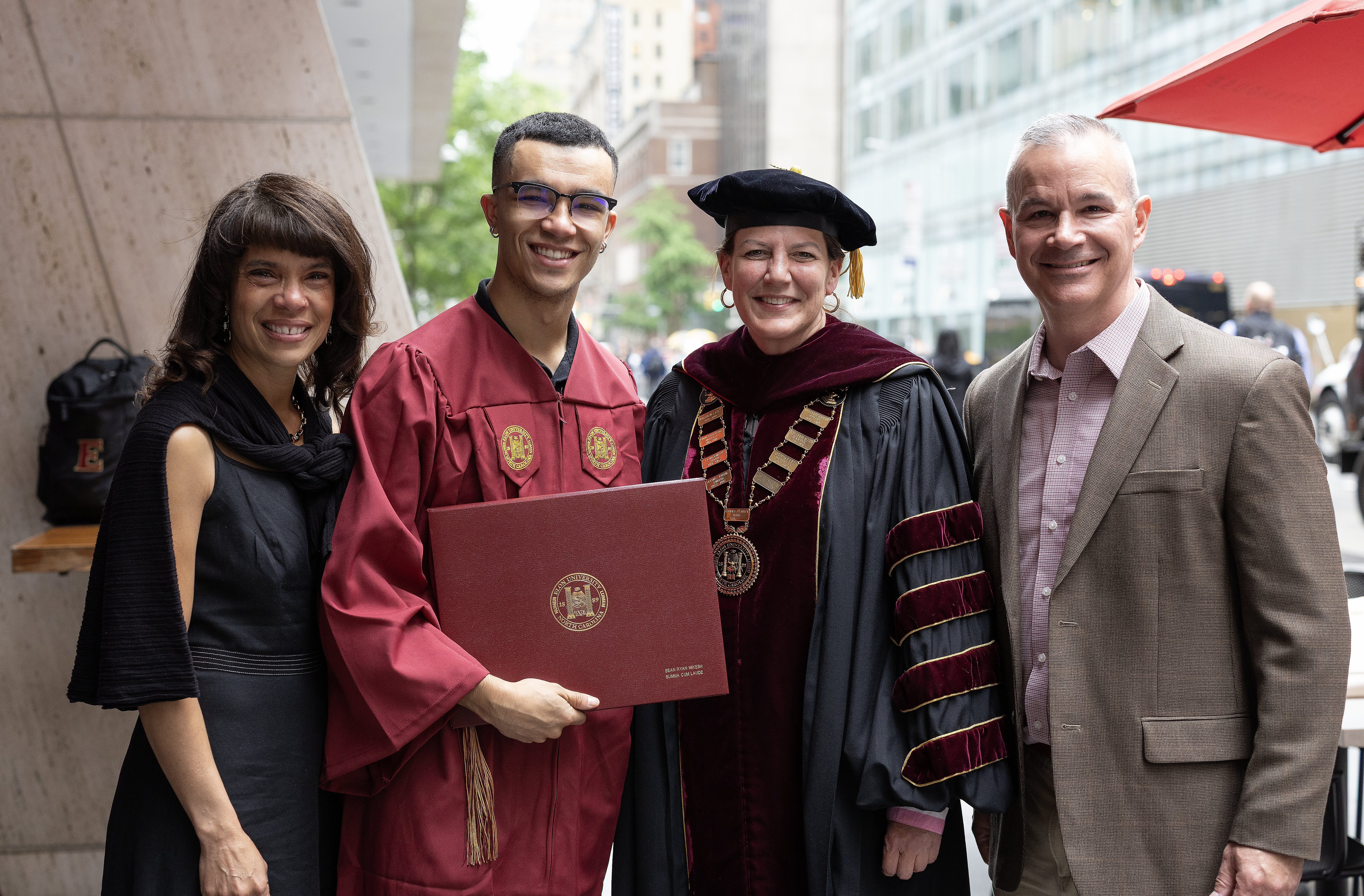 The graduate, holding his diploma, poses with the university president and two others, all smiling for a celebratory photo.