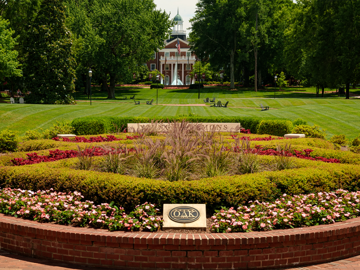 This photo depicts a beautifully manicured college campus, specifically Elon University in North Carolina. The image is taken from a symmetrical angle, showcasing: A lush, landscaped garden in the foreground, featuring pink flowers, ornamental grasses, and vibrant red and green shrubbery. A stone plaque in the center foreground that reads "OΔK", representing Omicron Delta Kappa, a national leadership honor society. Behind the garden, a sign engraved with "Alamance"—likely referring to Alamance Building, a prominent academic structure on Elon's campus. The Alamance Building is visible in the background, marked by its classic red-brick Georgian architecture, white columns, and a green-domed cupola. The surrounding quad area features perfectly mowed grass, park benches, walking paths, and mature trees, all contributing to a serene and collegiate atmosphere. This scene captures a quintessential view of Elon University’s campus—neatly arranged, vibrant, and steeped in academic tradition.