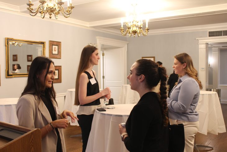 Several women engage in conversation around tall tables at a professional networking event in a warmly lit room.
