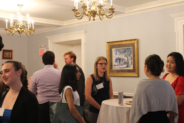 Groups of attendees chat and mingle around tall tables in a chandelier-lit room during a professional social gathering.