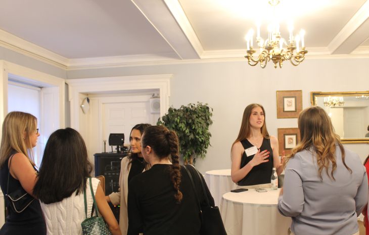 A young woman speaks to a small group during a networking event, while others converse nearby in a formal setting.