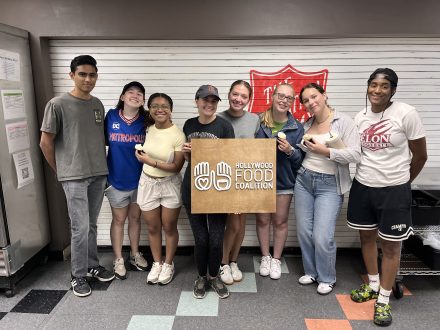 A group of eight volunteers smiles together indoors, holding a Hollywood Food Coalition sign in front of a service counter.