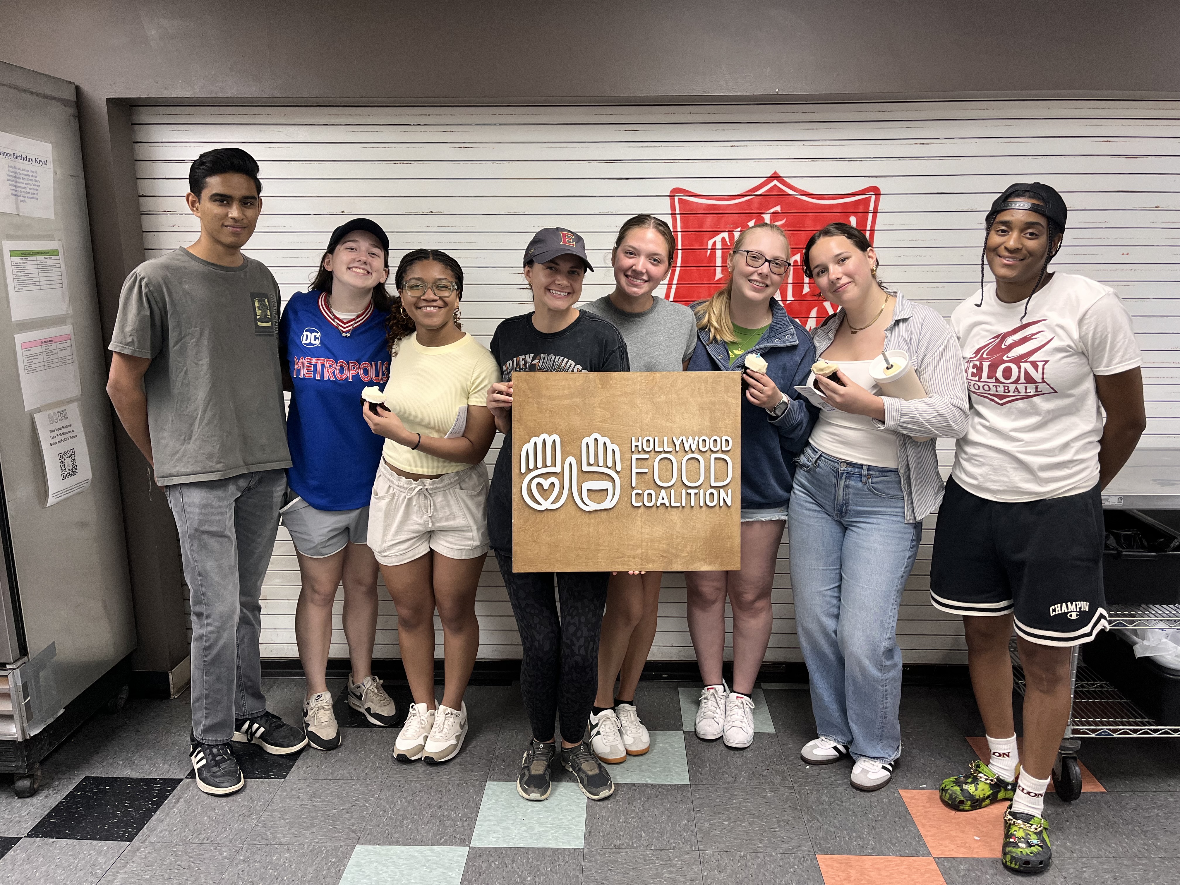 A group of eight volunteers smiles together indoors, holding a Hollywood Food Coalition sign in front of a service counter.