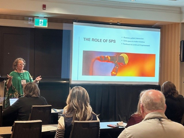 A woman presents to an audience in a conference room with a slide titled “The Role of SPS,” featuring a microphone image and bullet points about patient interaction, safe learning environments, and feedback.