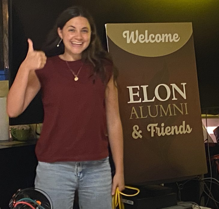 A woman gives a thumbs up while standing next to a sign that says “Welcome Elon Alumni & Friends.”