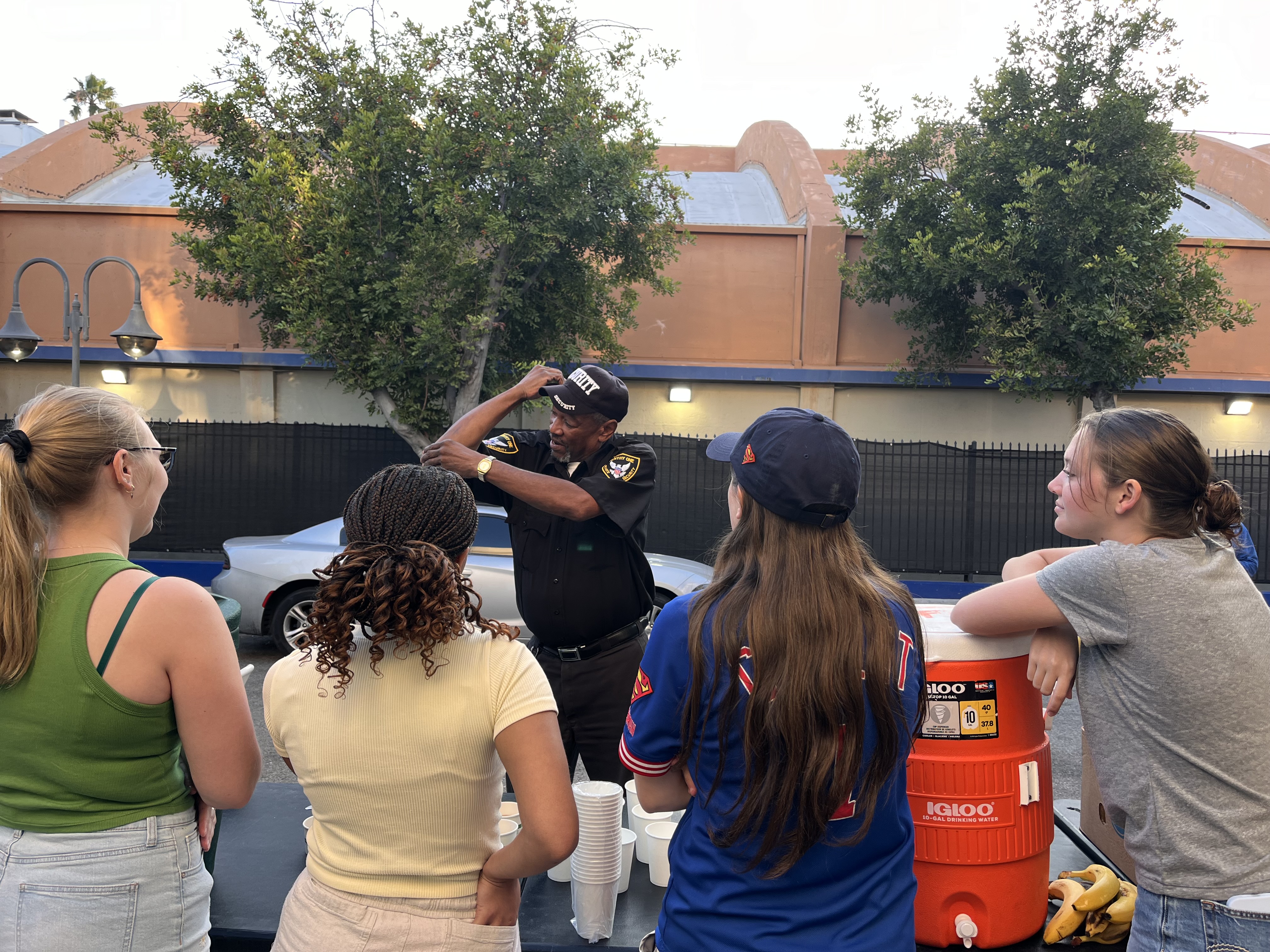 A security officer playfully flexes his arm while chatting with four smiling volunteers gathered around a drink station outdoors.
