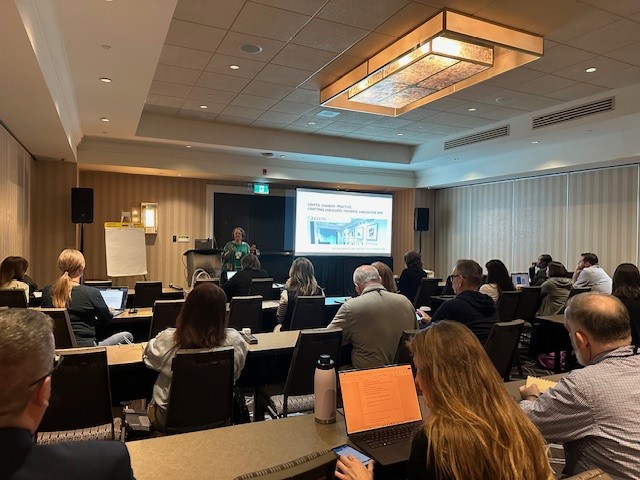A group of attendees listen to a presenter speaking in a conference room, with a slide projected at the front titled “SAFETY, HUMAN INTERACTION, & LEARNING ENVIRONMENTS.”