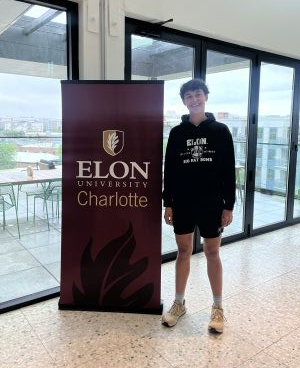 A student in an Elon sweatshirt smiles next to an “Elon University Charlotte” banner in a bright indoor space with large window