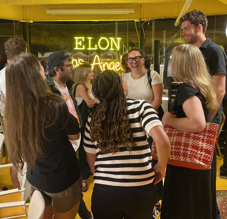 A group of smiling people enjoy a lively conversation under a glowing “Elon Los Angeles” sign.