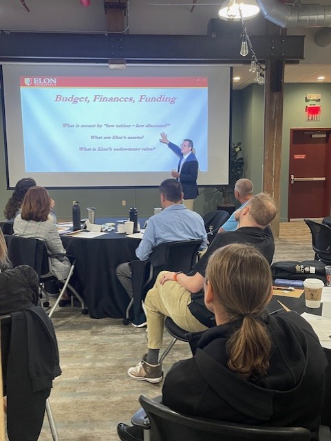 A speaker presents to an attentive audience, pointing at a slide titled “Budget, Finances, Funding” with discussion questions projected on the screen.