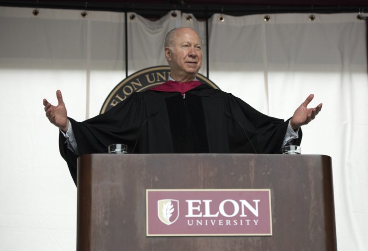 A man in an academic hood speaks behind an Elon University podium. His arms are outstreched as he gestures.