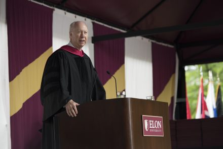 A man in an academic robe speaks behind a podium that says Elon University.