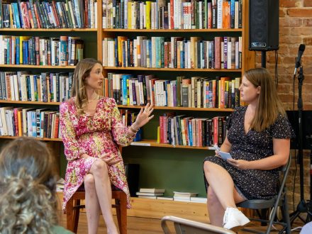 Two women seated in chairs facing an audience inside a bookstore. Shelves of books are seen behind them.