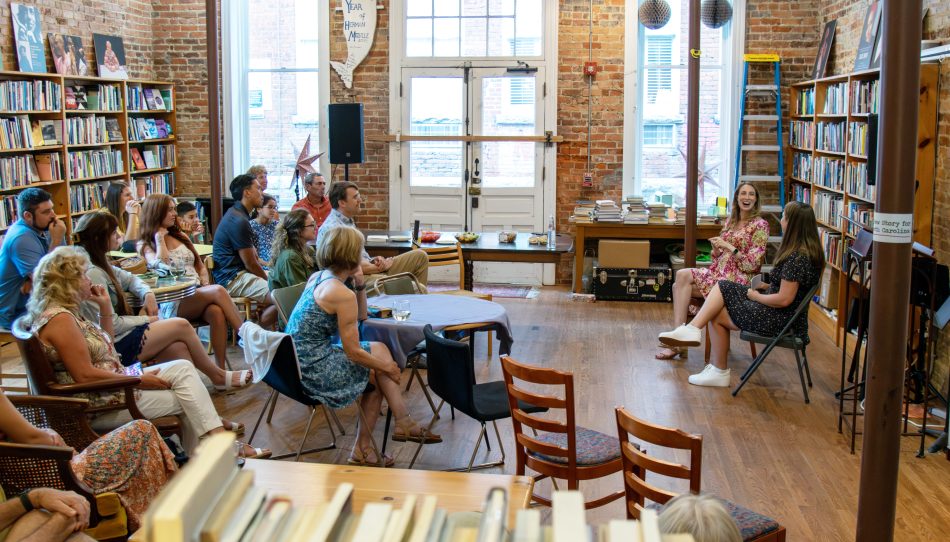 The back of a bookstore with people seated in chairs. Two women seated in front speak animatedly. Bookshelves are seen along the walls