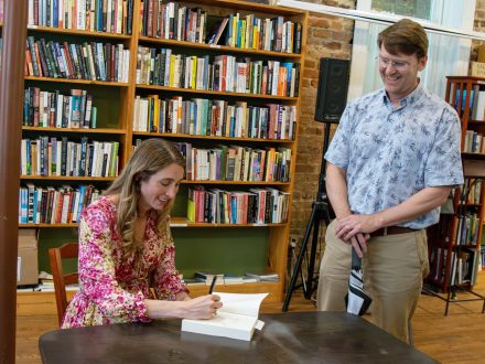 A woman at a table signs a book. A man stands by smiling.