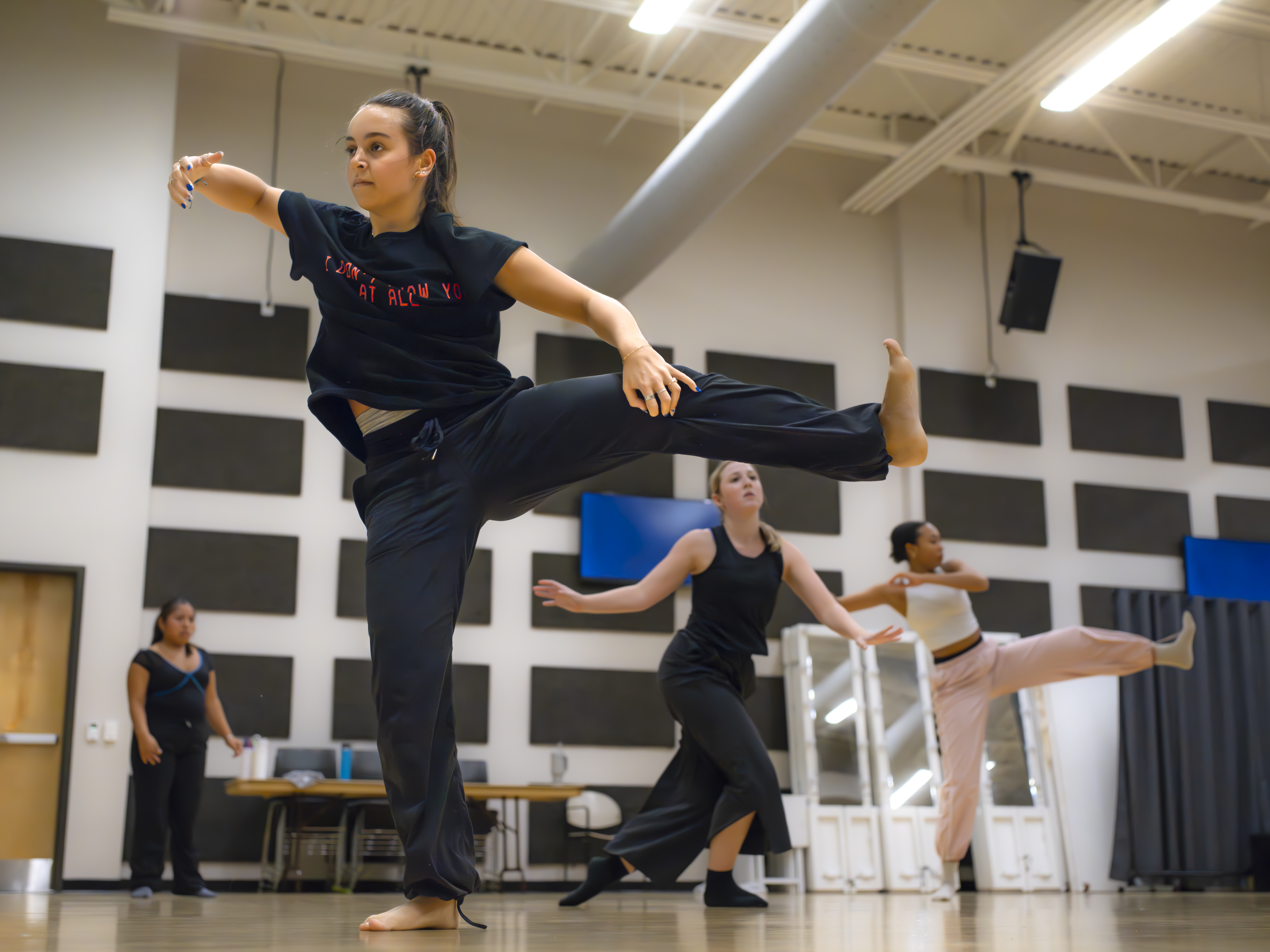 A group of dancers rehearses in a studio, with the lead dancer in front performing a high leg lift with focus and control.