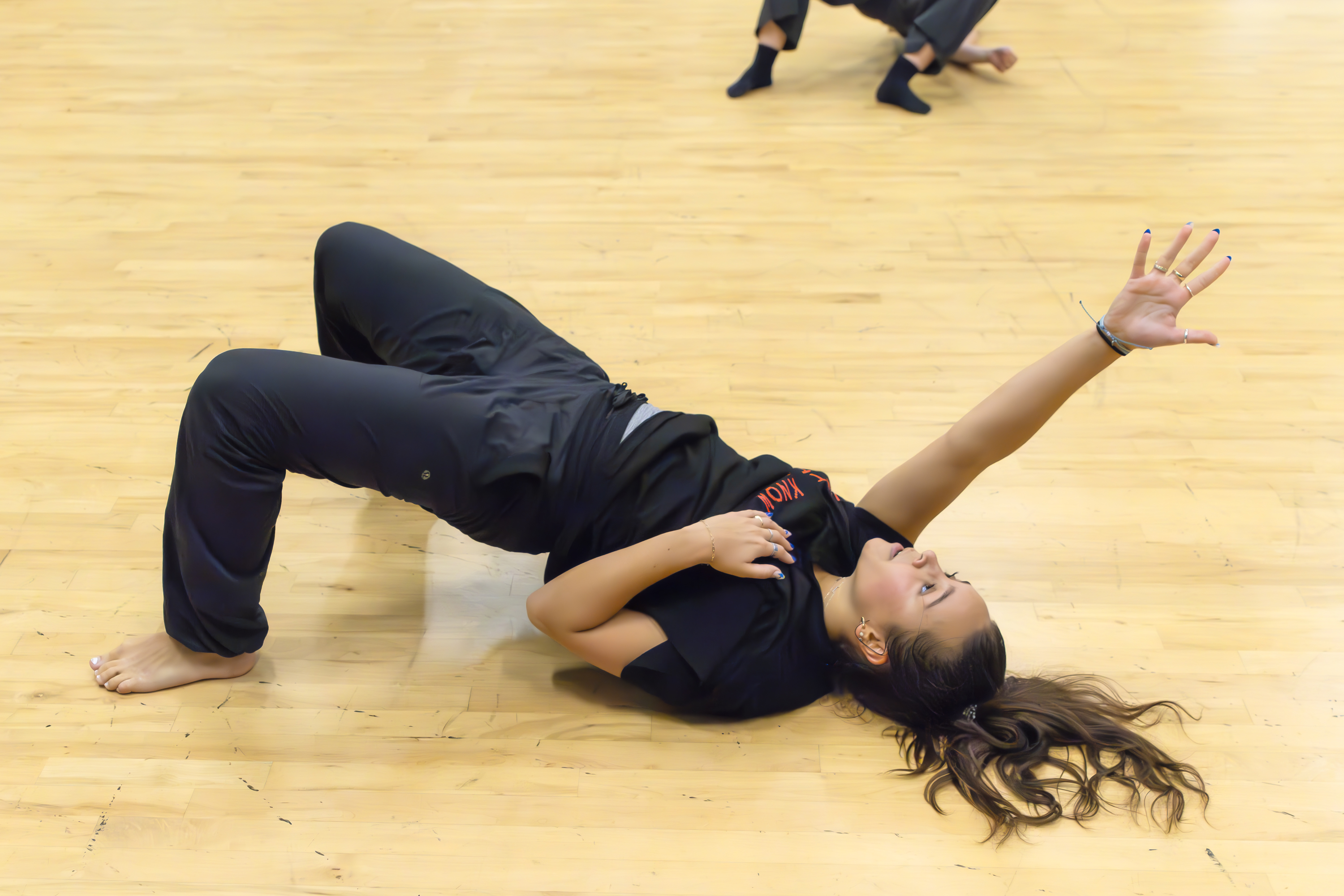 A dancer lies on the studio floor in a dramatic pose, arching her hips upward while extending one arm gracefully toward the ceiling.