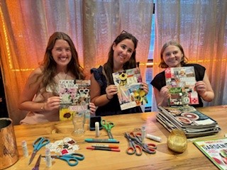 Three people smile while holding up their vision boards at the WE Envision event, seated at a craft table with scissors, markers, and magazines.
