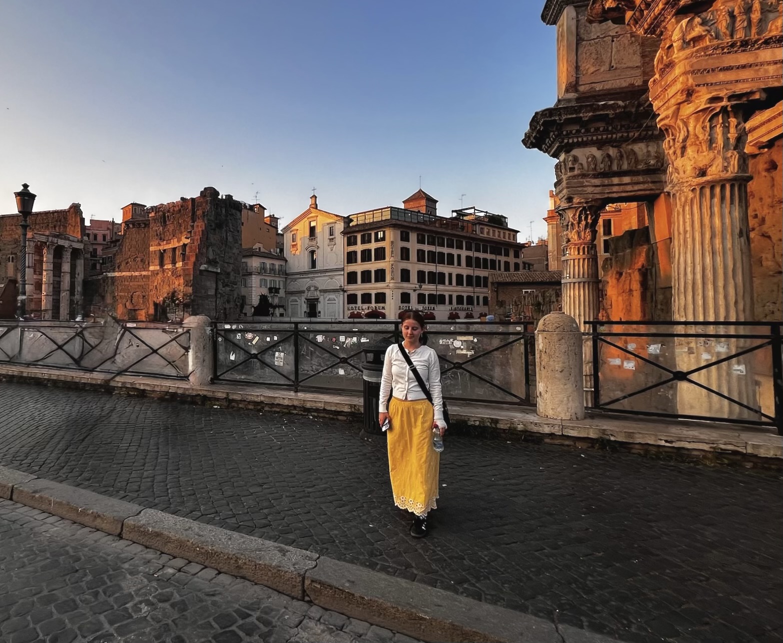 A young woman in a yellow skirt walks along a cobblestone street at sunset near ancient Roman ruins, with golden light illuminating the historic architecture around her.
